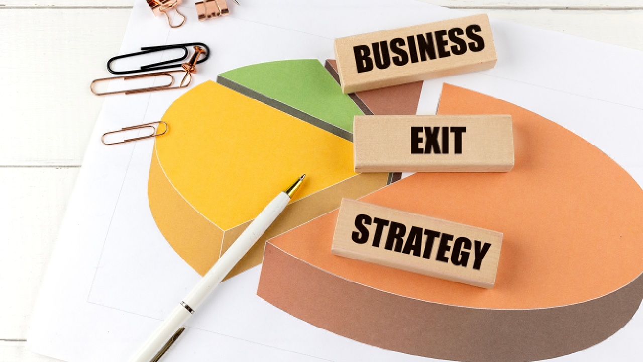 Illustration of a pie chart with coloured segments on a desk, alongside wooden blocks labelled “Business”, “Exit”, and “Strategy”, representing business exit strategy planning.