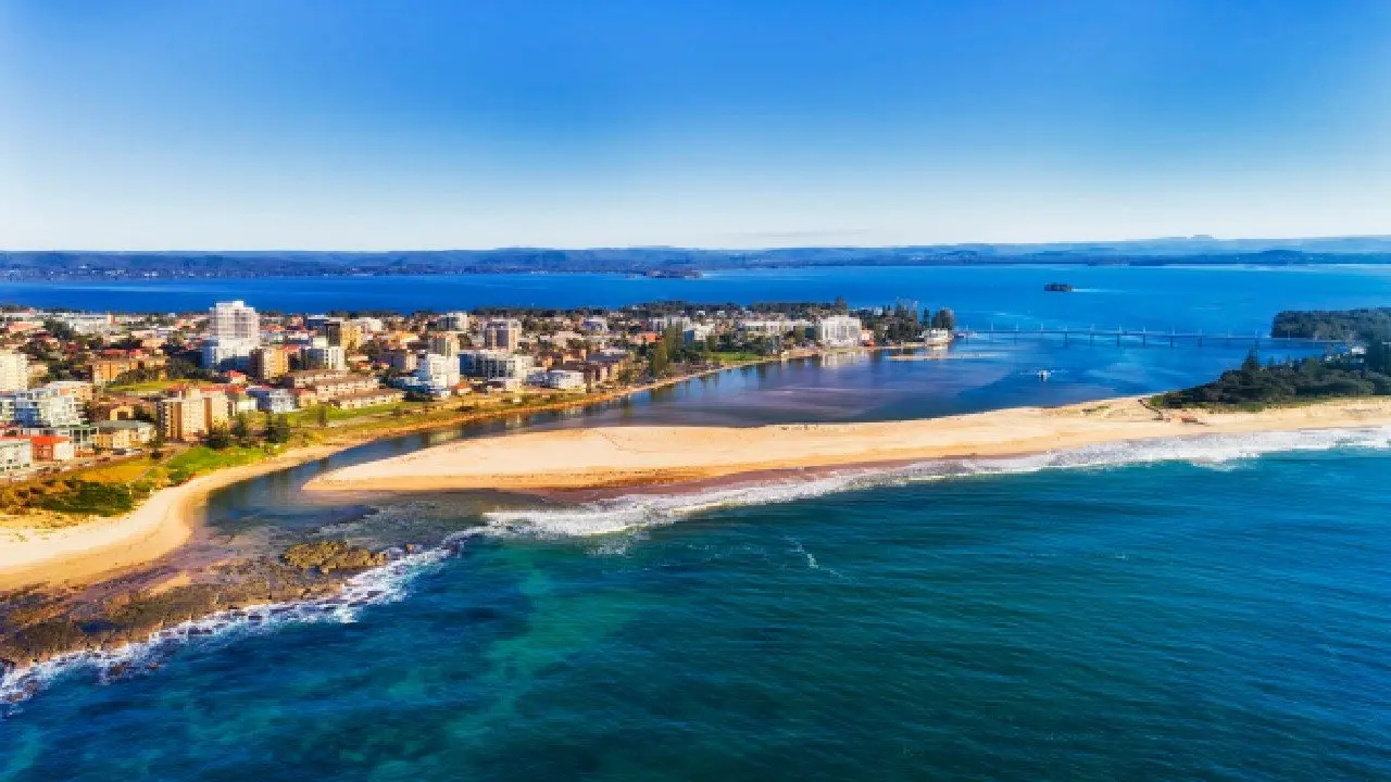 Aerial view of The Entrance on the Central Coast, showing coastal waterways, beach, and a vibrant strip of local businesses and residential buildings along the shoreline.